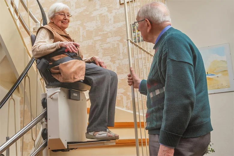 stair-lifts-780 An older woman sitting in a stairlift with her husband looking on.